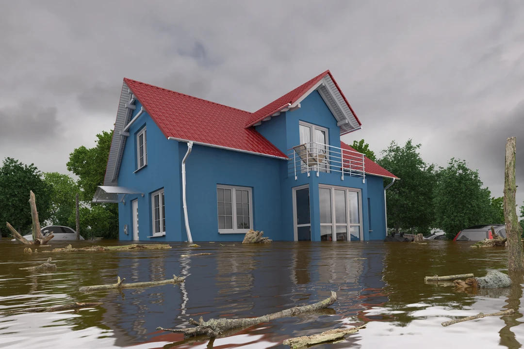 A blue house with a red roof partially submerged in murky floodwater filled with floating debris under a cloudy sky