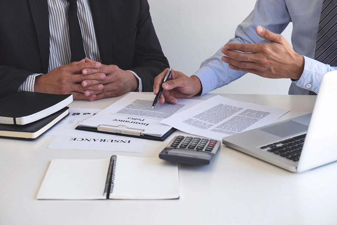 Two professionals in suits review and point to an insurance policy document on a desk with a laptop, calculator, and notebooks